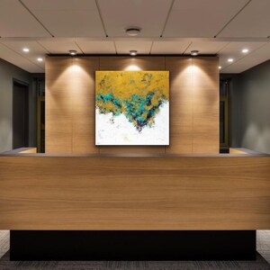 May include: A modern reception desk with a square abstract painting. The painting features gold, teal, and white colors. The desk is made of wood and has a black base. The room has recessed lighting and gray walls.