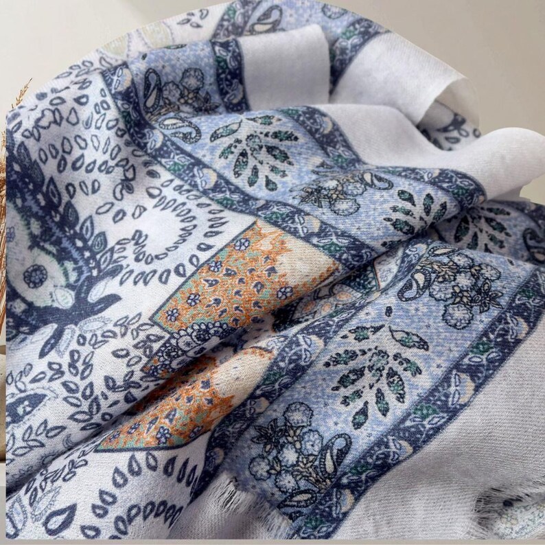 Op de afbeelding: Een gevouwen sjaal met een patchwork ontwerp. De sjaal heeft een mix van witte, blauwe en oranje patronen, waaronder bloemen- en paisley motieven. De randen hebben franjes.