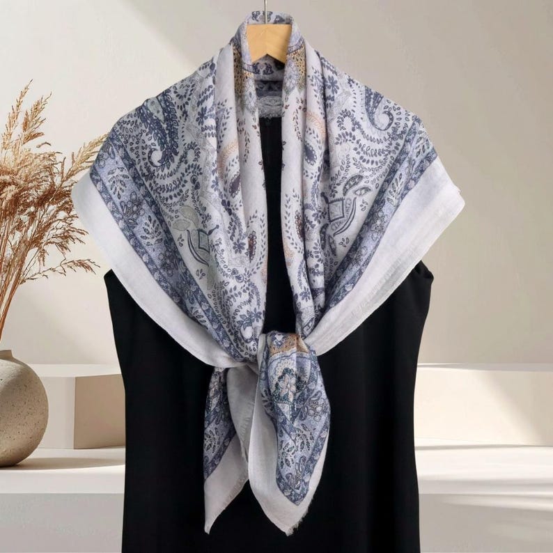 Op de afbeelding: Een vierkante sjaal met een paisley patroon in blauw, wit en beige tinten. De sjaal is gedrapeerd over een zwart kledingstuk, waarbij het ontwerp en de textuur worden benadrukt. De sjaal is aan de voorkant geknoopt.