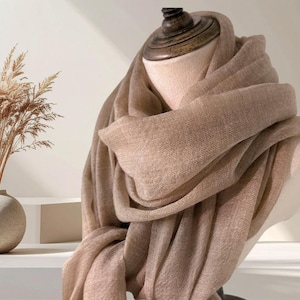 May include: A soft, light brown scarf draped elegantly around a mannequin neck. The fabric features a fine, woven texture with a subtle sheen, creating multiple folds and layers that showcase the neutral tan color and the lightweight material.