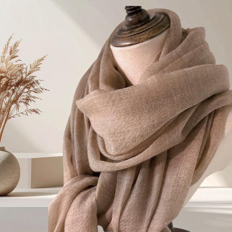 May include: A soft, light brown scarf draped elegantly around a mannequin neck. The fabric features a fine, woven texture with a subtle sheen, creating multiple folds and layers that showcase the neutral tan color and the lightweight material.
