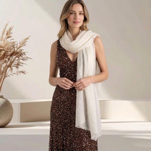 May include: A sleeveless, floor-length gown covered in dark brown sequins with a deep V-neckline, paired with a long, rectangular, off-white scarf featuring a soft, fringed edge that drapes over the shoulders and down the front of the dress.