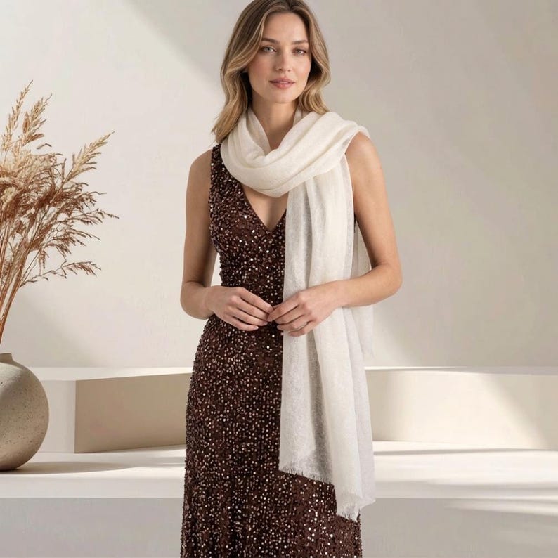 May include: A sleeveless, floor-length gown covered in dark brown sequins with a deep V-neckline, paired with a long, rectangular, off-white scarf featuring a soft, fringed edge that drapes over the shoulders and down the front of the dress.