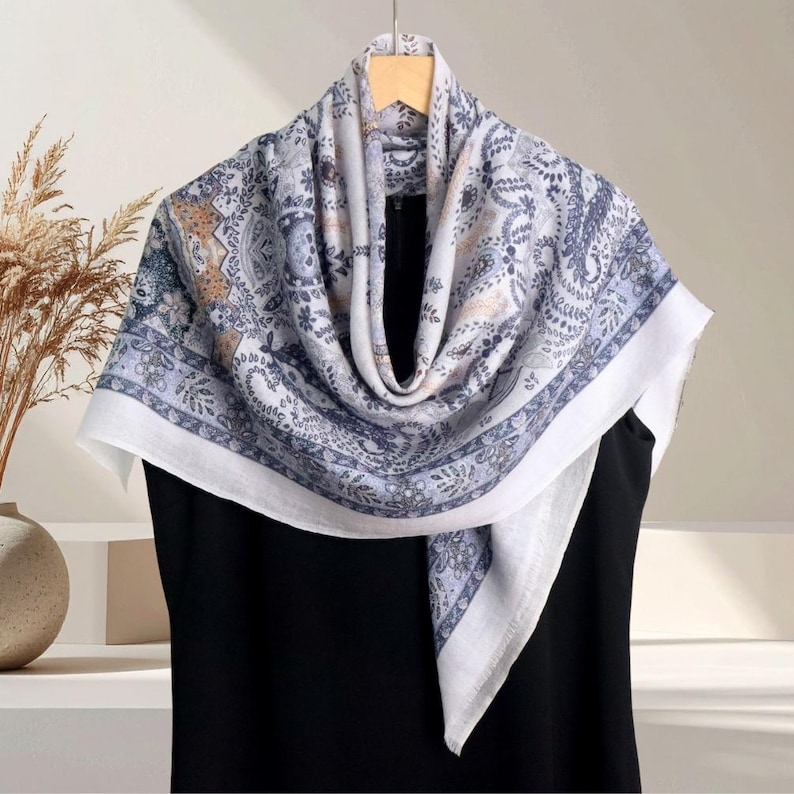 Op de afbeelding: Een paisley-patroon sjaal in blauw, wit en bruin, gedrapeerd over een zwart kledingstuk. De sjaal heeft een gedetailleerd bloemmotief en een witte rand. De sjaal hangt aan een houten hanger.