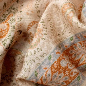 Op de afbeelding: Cr&egrave;mekleurige sjaal met ingewikkelde paisley- en bloempatronen in tinten oranje, groen en blauw. De sjaal heeft een gedetailleerde rand met geometrische ontwerpen. De stof lijkt zacht en vloeiend, met zichtbare textuur.