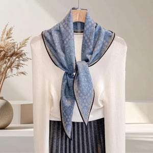 Puede incluir: Una bufanda azul claro con un estampado de cachemira y ribete negro está drapeada sobre un suéter blanco. La bufanda está anudada en la parte delantera, creando un accesorio elegante. La bufanda es de material suave.