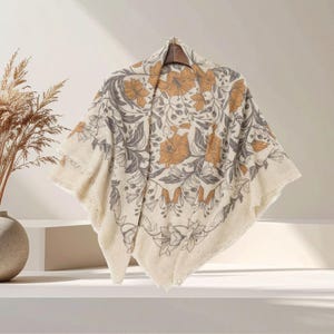 May include: A cream-colored square scarf featuring a repeating botanical pattern of muted orange flowers and gray foliage. The fabric has a soft, textured appearance with frayed edges, draped over a hanger to display the floral design clearly.