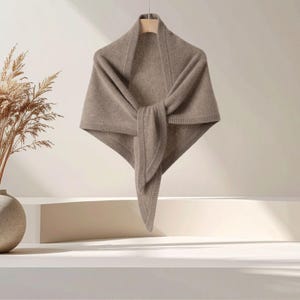 May include: A triangular taupe shawl featuring a soft knit texture and a ribbed border edge. The garment is draped in a pointed shape, showcasing a solid neutral color and a consistent woven pattern throughout the fabric construction.