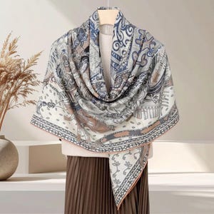May include: A large square scarf featuring a complex pattern of blue, brown, and cream tones. The design includes intricate paisley swirls, geometric borders, and stylized floral motifs. The scarf is draped over a light-colored top and a brown skirt.