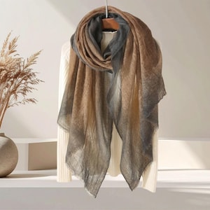 May include: A draped scarf with a gradient color scheme, transitioning from brown to gray. The scarf is displayed on a hanger, showcasing its soft, flowing fabric. The scarf's design features a fringed edge.