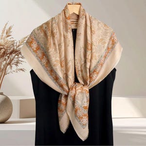 Op de afbeelding: Een beige sjaal met een ingewikkeld bloemen- en paisley-patroon in tinten oranje, blauw en bruin. De sjaal is over een zwart kledingstuk gedrapeerd en aan de voorkant geknoopt, waardoor het ontwerp wordt benadrukt. Het wordt gepresenteerd op een houten hanger.