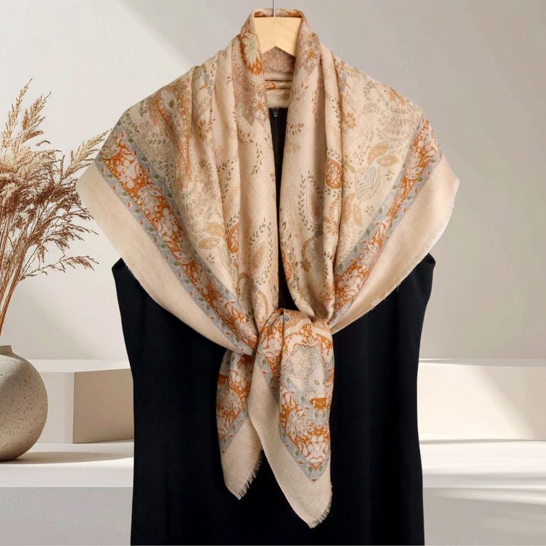 Op de afbeelding: Een beige sjaal met een ingewikkeld bloemen- en paisley-patroon in tinten oranje, blauw en bruin. De sjaal is over een zwart kledingstuk gedrapeerd en aan de voorkant geknoopt, waardoor het ontwerp wordt benadrukt. Het wordt gepresenteerd op een houten hanger.