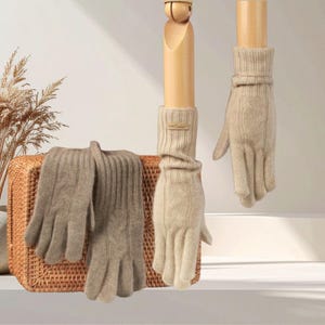 Könnte beinhalten: Ein Paar beigefarbene Strickhandschuhe mit Zopfmuster und gerippten Bündchen, die am Handgelenk mit einem kleinen rechteckigen Etikett versehen sind. Ein Handschuh wird auf einem Holzständer präsentiert, der andere liegt gefaltet auf einem Korb.