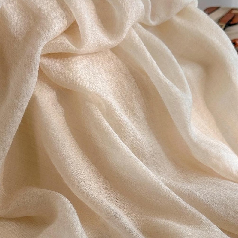 May include: A lightweight, semi-sheer scarf in a soft cream or off-white color. The fabric features a fine, slightly textured weave with visible folds and drapes that create subtle shadows and highlights across the surface of the material.