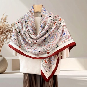 May include: A white scarf with a paisley pattern in shades of red, blue, green, and gold. The scarf has a white border with a red trim. The scarf is draped over a white sweater.