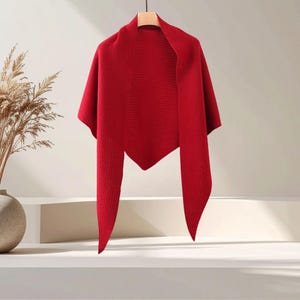 Puede incluir: Un chal triangular de color rojo vibrante con un patrón de tejido fino y texturizado. La prenda se muestra colgada de una percha, destacando su dobladillo en punta y silueta drapeada, con un color rojo sólido y uniforme en toda la pieza.