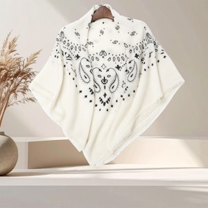 May include: A cream-colored poncho featuring a black paisley pattern printed across the fabric. The garment has a wide, draped silhouette with a pointed hemline and is displayed on a wooden hanger, showcasing the intricate black design details.