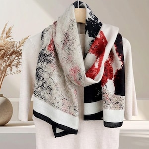 Peut inclure: Écharpe blanche avec un motif abstrait noir et rouge. L'écharpe est drapée sur un pull de couleur claire sur un cintre en bois. L'écharpe a une bordure noire.