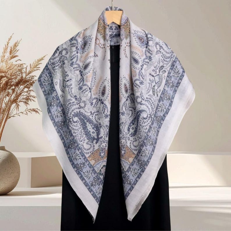 Op de afbeelding: Een vierkante sjaal met een paisley patroon in tinten wit, blauw en bruin. De sjaal is gedrapeerd over een zwart kledingstuk, waarbij het ingewikkelde ontwerp en de rand worden getoond. De sjaal is gemaakt van een zacht, lichtgewicht materiaal.