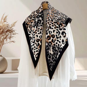 Puede incluir: Un pañuelo cuadrado con estampado de leopardo en tonos marrón, negro y gris. El pañuelo tiene un borde negro y está drapeado sobre una camisa blanca. El pañuelo está hecho de un material suave y ligero.