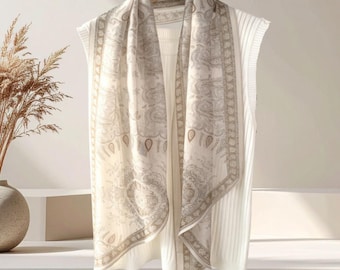 May include: A cream-coloured scarf with a paisley pattern in beige and brown tones is draped over a white ribbed sweater. The scarf hangs on a wooden hanger, creating a layered look. The overall aesthetic is soft and elegant.
