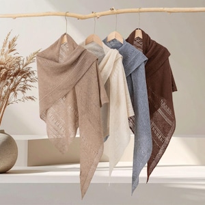 May include: Four triangular knit shawls hanging on wooden hangers. The shawls are displayed in a row, featuring colours of light brown, cream, dusty blue, and dark brown. Each shawl has a delicate, openwork pattern with vertical stripes.