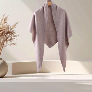 May include: A triangular shawl in a muted mauve colour, featuring a fine-knit texture with a subtle open-work pattern along the centre panel. The garment is draped over a wooden hanger, displaying its pointed hem and soft, lightweight fabric construction.