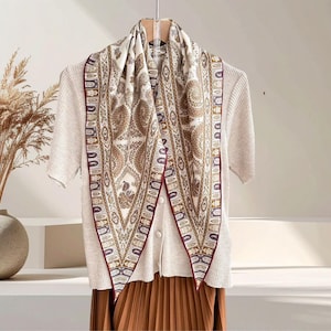 May include: A cream scarf featuring a paisley design in brown and gold, edged with a burgundy border. The scarf is draped over a light-coloured sweater and a pleated brown skirt, creating a layered appearance.