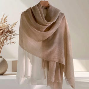 May include: A light beige, rectangular scarf with a fine, woven texture and frayed edges, draped over a plain white long-sleeved shirt. The scarf features a soft, neutral color palette and is displayed on a hanger against a neutral background.