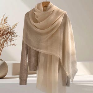May include: A lightweight, semi-sheer beige scarf draped over a long-sleeved, light brown knit sweater. The scarf features a soft, fine texture and hangs in loose, flowing folds, while the sweater has a ribbed hem and cuffs in a solid tone.