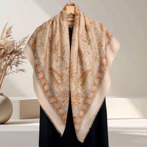 Op de afbeelding: Een beige paisley-patroon sjaal met oranje, blauwe en grijze accenten. De sjaal is over een houten hanger gedrapeerd, waarbij de vierkante vorm en de gefranjerde randen worden getoond. Het ontwerp heeft een gedetailleerd bloemenpatroon.