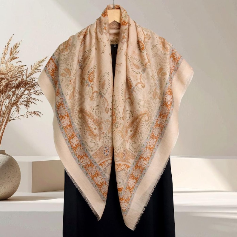 Op de afbeelding: Een beige paisley-patroon sjaal met oranje, blauwe en grijze accenten. De sjaal is over een houten hanger gedrapeerd, waarbij de vierkante vorm en de gefranjerde randen worden getoond. Het ontwerp heeft een gedetailleerd bloemenpatroon.