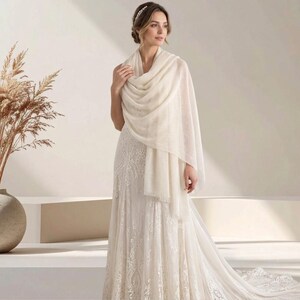 May include: A long, cream-colored rectangular shawl with a soft, lightweight texture and fringed edges, draped elegantly over the shoulders of a floor-length white lace gown featuring intricate floral embroidery and a flowing, full-length skirt.