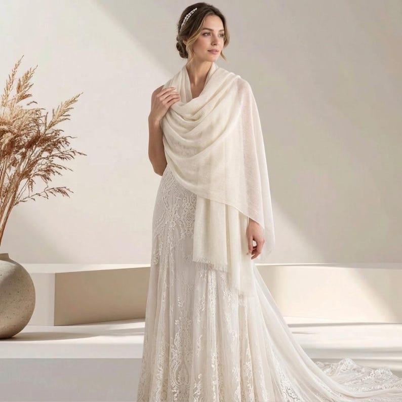 May include: A long, cream-colored rectangular shawl with a soft, lightweight texture and fringed edges, draped elegantly over the shoulders of a floor-length white lace gown featuring intricate floral embroidery and a flowing, full-length skirt.