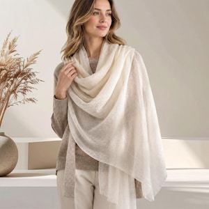 May include: A lightweight, off-white, semi-sheer rectangular scarf with a fine, delicate knit texture. The scarf is draped loosely over the shoulders, featuring soft, unfinished edges and a long, flowing silhouette that hangs down the front.