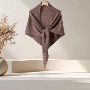 May include: A triangular knit shawl in a solid medium brown color. The garment features a ribbed border along the top edge and is draped to form a point at the center. The texture appears soft and consistent throughout the entire piece.