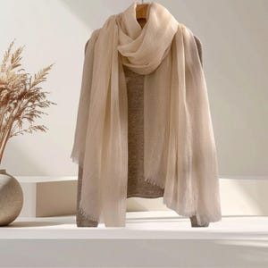 May include: A lightweight, cream-colored scarf with a soft, semi-sheer texture and delicate fringed edges, draped elegantly over a matching long-sleeved, knit sweater. The scarf features a loose weave and a neutral, solid tone throughout the fabric.