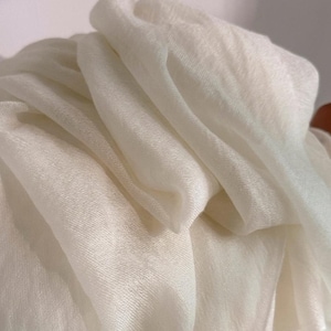 May include: A lightweight, off-white scarf with a soft, semi-sheer texture. The fabric is draped in loose, flowing folds, creating a series of gentle curves and shadows that highlight the fine, woven quality of the material throughout the piece.