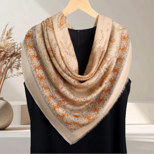 Op de afbeelding: Een beige vierkante sjaal met een ingewikkeld bloemen- en paisley patroon in oranje, groen en blauw. De sjaal is gedrapeerd over een zwart kledingstuk, waarbij het ontwerp en de gefranjerde randen worden getoond. De sjaal is gemaakt van een zacht materiaal.