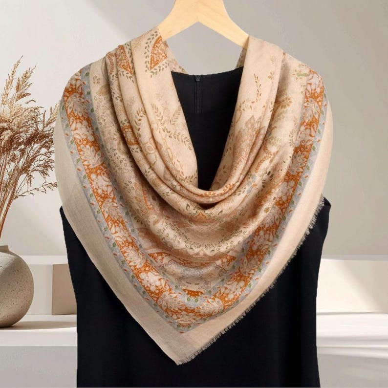 Op de afbeelding: Een beige vierkante sjaal met een ingewikkeld bloemen- en paisley patroon in oranje, groen en blauw. De sjaal is gedrapeerd over een zwart kledingstuk, waarbij het ontwerp en de gefranjerde randen worden getoond. De sjaal is gemaakt van een zacht materiaal.