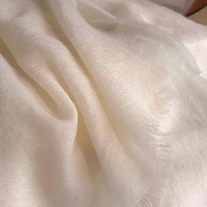 May include: A solid white scarf featuring a lightweight, semi-sheer fabric with a fine, woven texture. The edges of the scarf are finished with a delicate, frayed fringe detail, creating a soft and flowing appearance with natural folds.