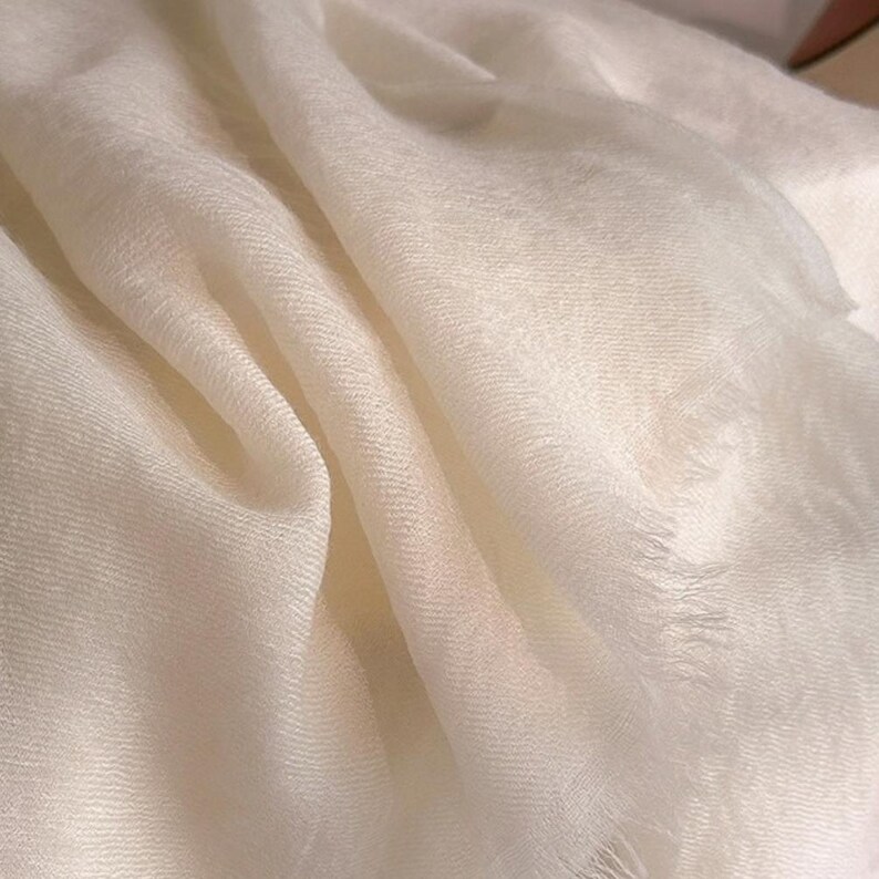 May include: A solid white scarf featuring a lightweight, semi-sheer fabric with a fine, woven texture. The edges of the scarf are finished with a delicate, frayed fringe detail, creating a soft and flowing appearance with natural folds.