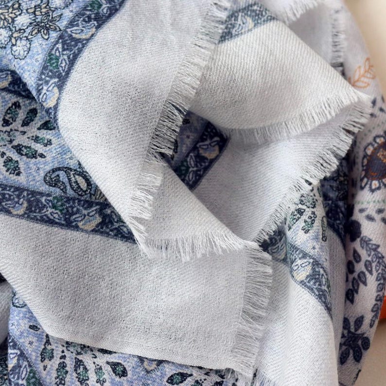 Op de afbeelding: Close-up van een gevouwen witte sjaal met een gedetailleerd paisley- en bloemenpatroon in tinten blauw, groen en grijs. De sjaal heeft een gefranjerde rand, wat textuur toevoegt. De stof lijkt zacht en licht.