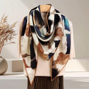 May include: A square scarf featuring an abstract pattern of cream, black, brown, and teal blue shapes. The scarf has a thin brown border along its edges and is draped over a light-coloured top, displaying a smooth texture and a folded design.
