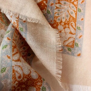 Op de afbeelding: Close-up van een beige sjaal met een gefranjerde rand. De sjaal heeft een herhalend patroon van oranje en blauwe paisley-ontwerpen. De stof is gevouwen en onthult de textuur en kleurvariaties.