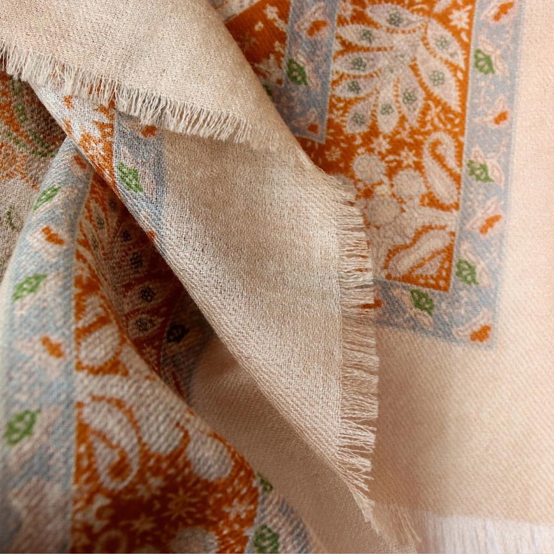 Op de afbeelding: Close-up van een beige sjaal met een gefranjerde rand. De sjaal heeft een herhalend patroon van oranje en blauwe paisley-ontwerpen. De stof is gevouwen en onthult de textuur en kleurvariaties.
