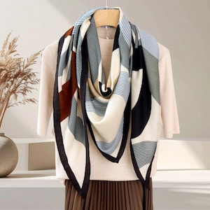 May include: A silk scarf with a geometric pattern in black, white, brown, and blue. The scarf is draped over a beige top and brown skirt. The scarf has a black border and is hanging on a wooden hanger.