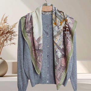 Peut inclure: Un cardigan gris avec une fermeture boutonnée est présenté sur un cintre en bois. Une écharpe en soie à motifs floraux et géométriques dans les tons de rose, vert et beige est drapée sur le cardigan.