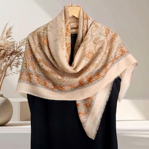 May include: A beige scarf with an intricate floral and paisley pattern in shades of orange, green, and grey. The scarf is draped over a black garment, showcasing its design and fringed edges. It hangs from a wooden hanger.