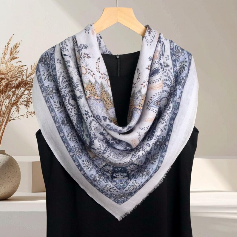 Op de afbeelding: Een vierkante sjaal met een gedetailleerd paisley patroon in tinten wit, blauw en bruin. De sjaal is over een zwart kledingstuk gedrapeerd en hangt aan een houten hanger. De randen van de sjaal hebben een gefranjerde afwerking.